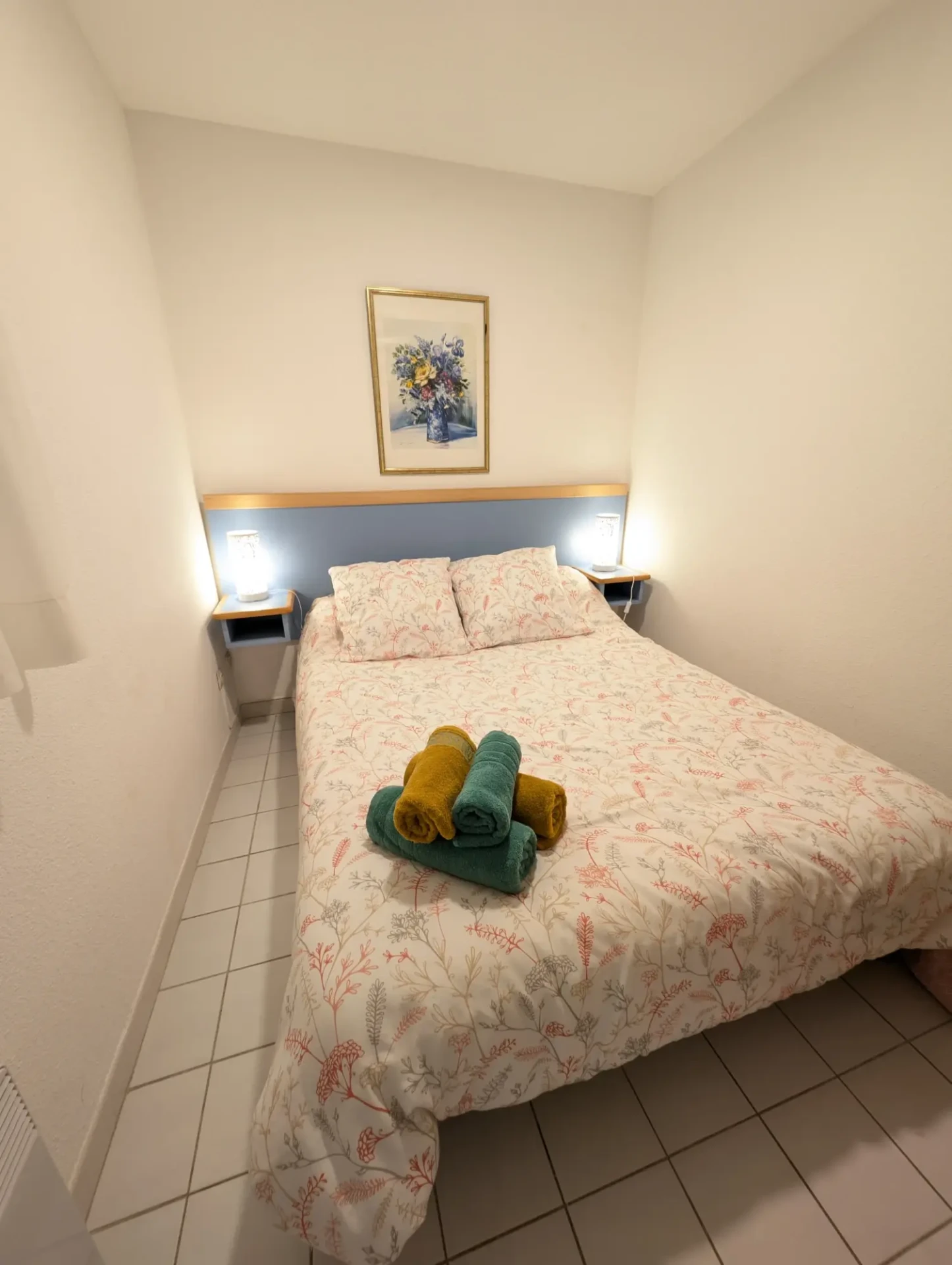 Chambre avec murs blancs, literie fleurie claire et serviettes vertes et jaunes